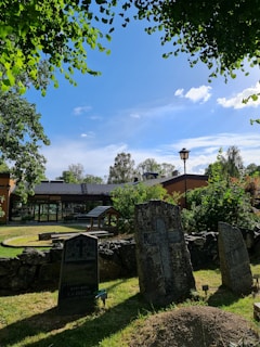 An outdoor scene featuring several old gravestones in a cemetery, surrounded by lush greenery and trees under a clear blue sky. There is a building with a modest, modern design in the background, and a well-maintained grass area with a stone wall in the foreground.