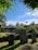 An outdoor scene featuring several old gravestones in a cemetery, surrounded by lush greenery and trees under a clear blue sky. There is a building with a modest, modern design in the background, and a well-maintained grass area with a stone wall in the foreground.
