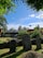 An outdoor scene featuring several old gravestones in a cemetery, surrounded by lush greenery and trees under a clear blue sky. There is a building with a modest, modern design in the background, and a well-maintained grass area with a stone wall in the foreground.