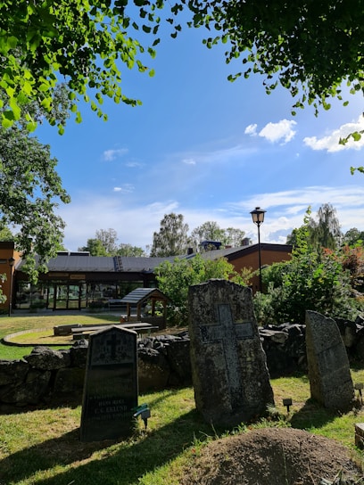 An outdoor scene featuring several old gravestones in a cemetery, surrounded by lush greenery and trees under a clear blue sky. There is a building with a modest, modern design in the background, and a well-maintained grass area with a stone wall in the foreground.