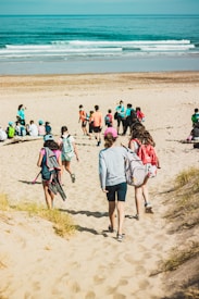 A group of people, mostly young, walking down a sandy path towards a beach. Some are carrying backpacks and wearing casual beach attire. The scene is sunny with the ocean visible in the background, featuring gentle waves and a clear blue sky.