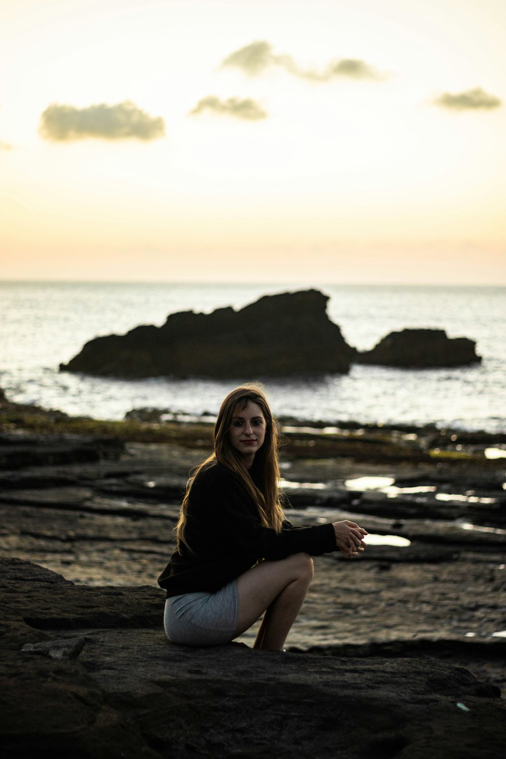 woman in black long sleeve shirt and black pants sitting on brown rock near sea during