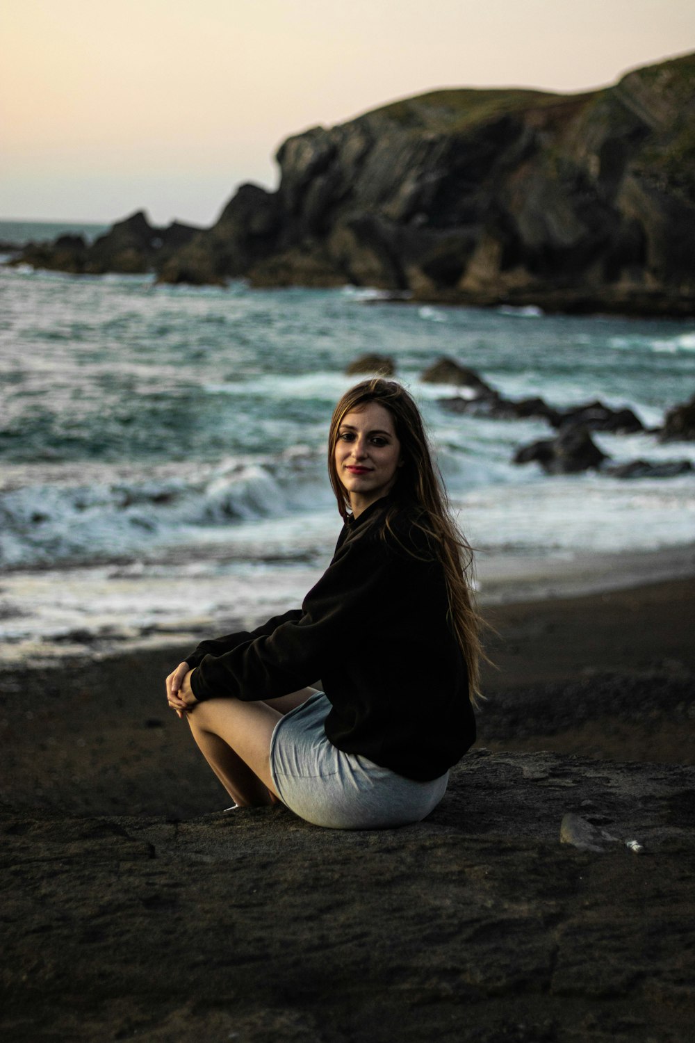 woman in black jacket sitting on beach shore during daytime