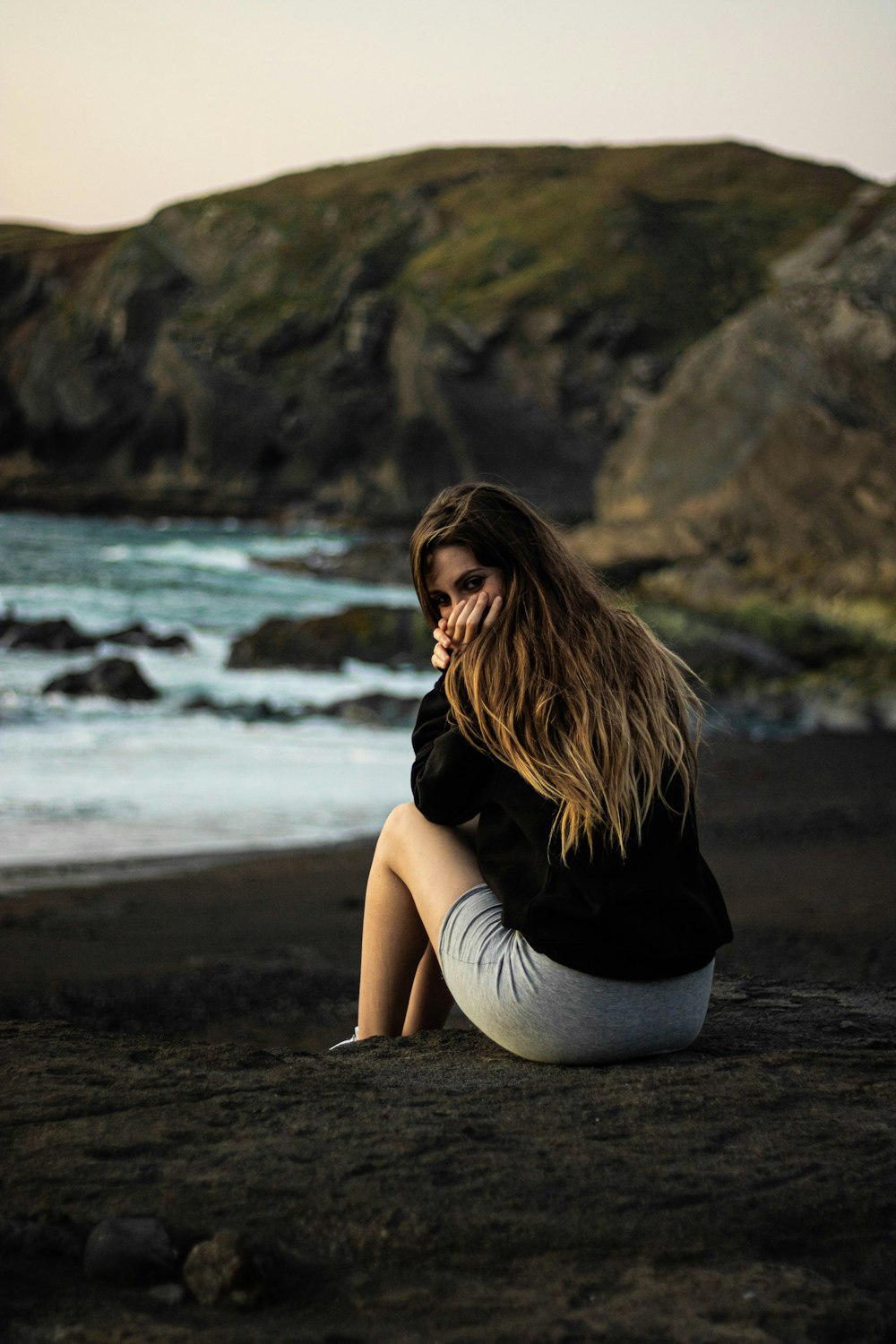 woman in black shirt and gray pants sitting on brown sand near body of water during