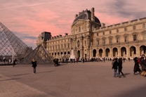 A historical museum building with ornate architecture stands prominently with a glass pyramid in the foreground. Numerous people are walking around or gathered in small groups on the broad plaza under a sky painted with soft pink hues.