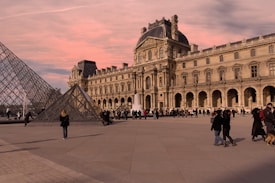 A historical museum building with ornate architecture stands prominently with a glass pyramid in the foreground. Numerous people are walking around or gathered in small groups on the broad plaza under a sky painted with soft pink hues.