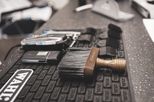 Various barbershop tools are neatly arranged on a countertop mat. A hairbrush with a wooden handle is prominently placed in the foreground, while behind it are several clipper guards and an electric hair clipper.