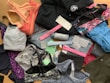 A collection of various athletic apparel and accessories laid out, including sports bras, tops, headbands, and tags. The items are in different colors and patterns, such as pastel pink, blue, and floral designs. A black jacket and several pieces still have brand tags attached.
