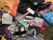 A collection of various athletic apparel and accessories laid out, including sports bras, tops, headbands, and tags. The items are in different colors and patterns, such as pastel pink, blue, and floral designs. A black jacket and several pieces still have brand tags attached.