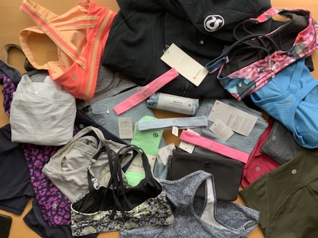 A collection of various athletic apparel and accessories laid out, including sports bras, tops, headbands, and tags. The items are in different colors and patterns, such as pastel pink, blue, and floral designs. A black jacket and several pieces still have brand tags attached.