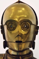 A humanoid robot with a shiny metallic gold surface and intricate detailing, including round eyes with grate-like designs and a small rectangular mouth. The head has a smooth, glossy finish with subtle reflections.
