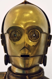 A humanoid robot with a shiny metallic gold surface and intricate detailing, including round eyes with grate-like designs and a small rectangular mouth. The head has a smooth, glossy finish with subtle reflections.
