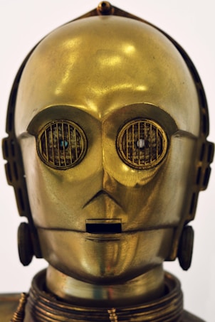 A humanoid robot with a shiny metallic gold surface and intricate detailing, including round eyes with grate-like designs and a small rectangular mouth. The head has a smooth, glossy finish with subtle reflections.
