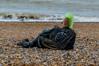 A person with a green mohawk and studded jacket is sitting on a pebble beach, facing the sea. The jacket displays various designs and patches, contributing to a punk aesthetic. The scene is set against a backdrop of overcast weather, with waves rolling onto the shore.