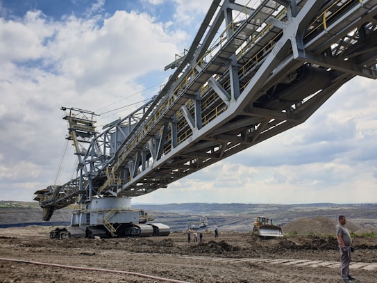A massive industrial machine, likely an excavator or conveyor, dominates a barren, open landscape. The imposing structure is made of metal, with intricate frameworks and large tracks for mobility. Several small figures of workers and a bulldozer can be seen nearby, giving a sense of scale. The sky is partly cloudy and expansive.