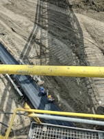 Two construction workers wearing safety helmets sit on a large conveyor belt system situated above a cleared dirt site. Shadows from the structure above create patterns on the ground. Yellow metal railings are visible in the foreground, indicating a viewing point or walkway above the belt.