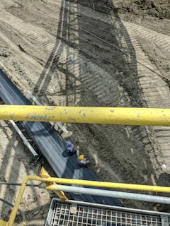 Two construction workers wearing safety helmets sit on a large conveyor belt system situated above a cleared dirt site. Shadows from the structure above create patterns on the ground. Yellow metal railings are visible in the foreground, indicating a viewing point or walkway above the belt.