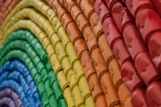 Colored barrels are stacked in an arched pattern, forming a visually striking arrangement with a spectrum of hues ranging from red and orange to yellow, green, blue, and purple. The barrels appear to be made of metal and are slightly dented, with some showing signs of wear.