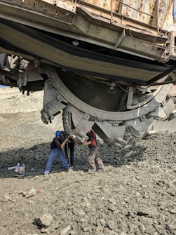Two workers interact with a large piece of industrial machinery, possibly for maintenance or repair. The ground is rough and composed of gravel or rocks, and various tools and containers are scattered nearby. The machine dominates the upper part of the image, appearing heavy and complex with a metallic construction.