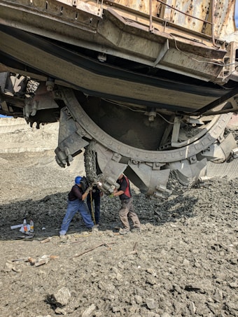 Two workers interact with a large piece of industrial machinery, possibly for maintenance or repair. The ground is rough and composed of gravel or rocks, and various tools and containers are scattered nearby. The machine dominates the upper part of the image, appearing heavy and complex with a metallic construction.