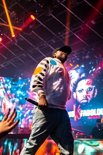 A person wearing a colorful, patterned jacket and cap is holding a microphone on stage. The background is vibrant with lights and a digital screen displaying graphics and text. The stage is illuminated with red and orange stage lights, the atmosphere appears energetic.