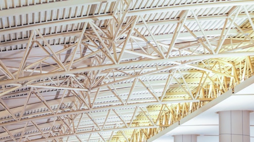 A detailed network of beige steel beams and trusses supporting the ceiling of a large building. The beams are geometrically arranged, creating a pattern across the ceiling. Columns, also beige, support the structure below.