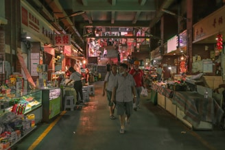 A bustling indoor market with various shops lining both sides of a central walkway. Shoppers walk through the dimly lit corridor, lined with colorful merchandise and illuminated signs. The upper floor is visible in the distance, accessed by a set of stairs, where more people can be seen in the background.