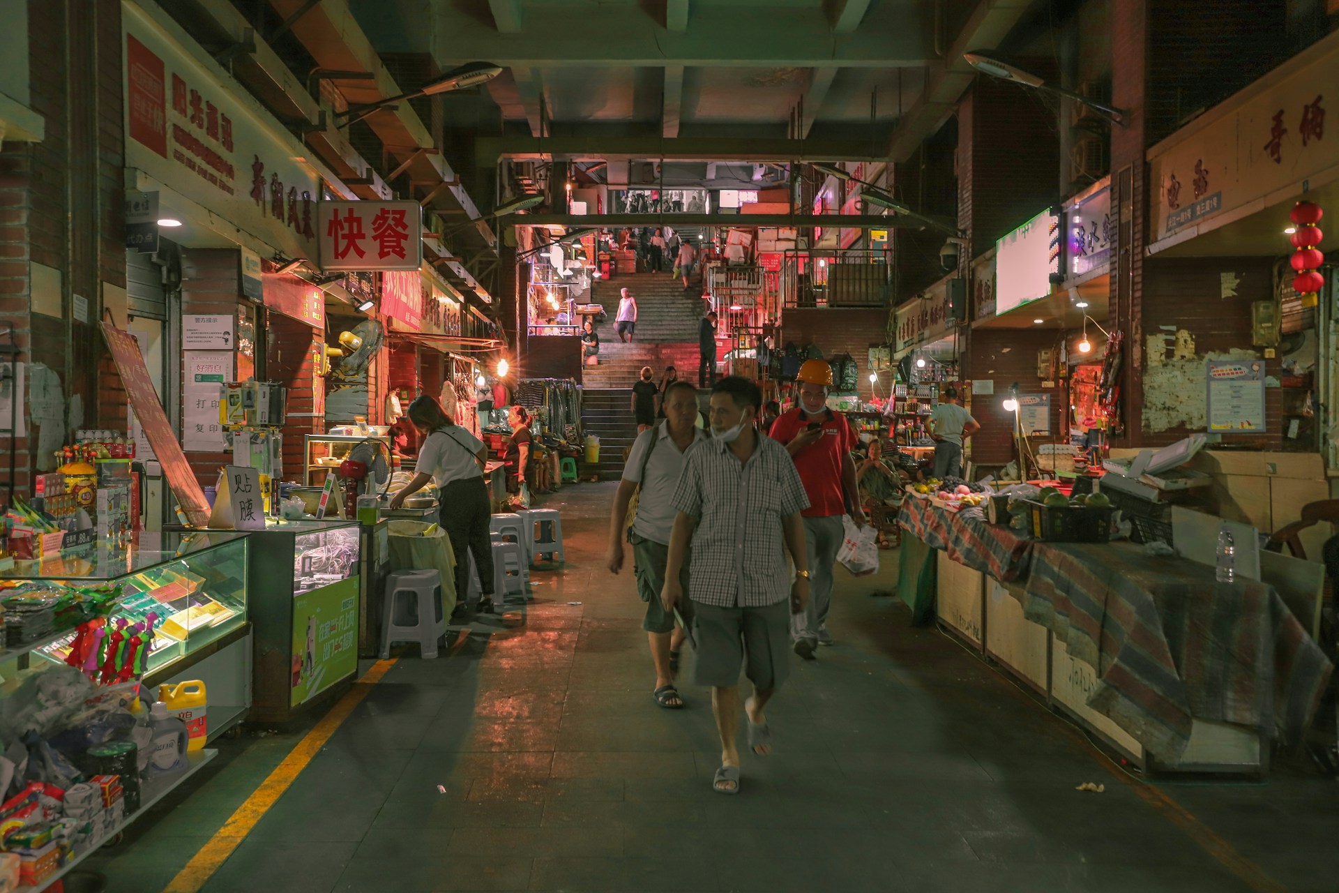 A bustling indoor market with various shops lining both sides of a central walkway. Shoppers walk through the dimly lit corridor, lined with colorful merchandise and illuminated signs. The upper floor is visible in the distance, accessed by a set of stairs, where more people can be seen in the background.