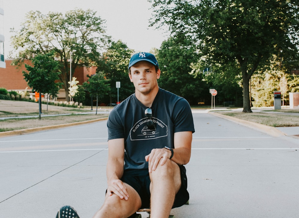 man in black crew neck t-shirt and blue fitted cap sitting on gray concrete pavement