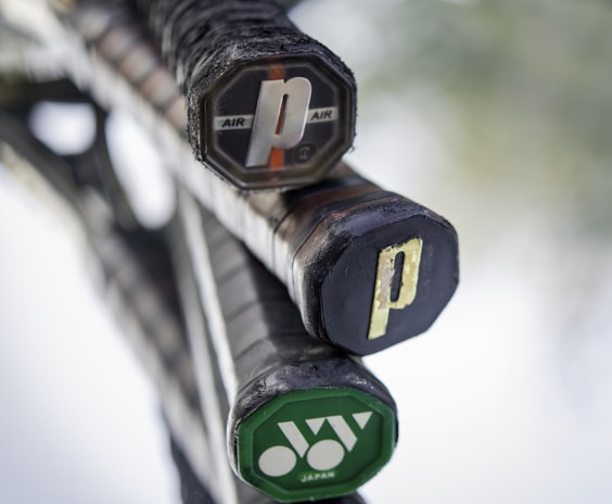 Close-up of the handles of three tennis rackets leaning together, with visible brand markings. The handles are black with textured grips, and each end cap has a unique design or logo, one being green and another featuring the word 'AIR'. The background is out of focus, highlighting the rackets.