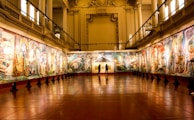 An art gallery with large, colorful murals adorning the walls. The room features high ceilings supported by decorative columns, and the lighting highlights the intricate details of the artwork. Three people stand in the center of the room, observing the art.