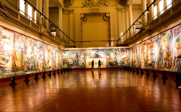 An art gallery with large, colorful murals adorning the walls. The room features high ceilings supported by decorative columns, and the lighting highlights the intricate details of the artwork. Three people stand in the center of the room, observing the art.