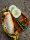 A serving of grilled rice wrapped in a banana leaf sits on a wooden platter. Accompanying the rice are two slices of cucumber, two long green beans, and a small portion of spicy chili paste with a few leaves.