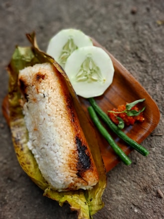 A serving of grilled rice wrapped in a banana leaf sits on a wooden platter. Accompanying the rice are two slices of cucumber, two long green beans, and a small portion of spicy chili paste with a few leaves.