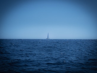 A lone sailboat cutting through deep blue ocean waves under a vast, open sky at sunset.