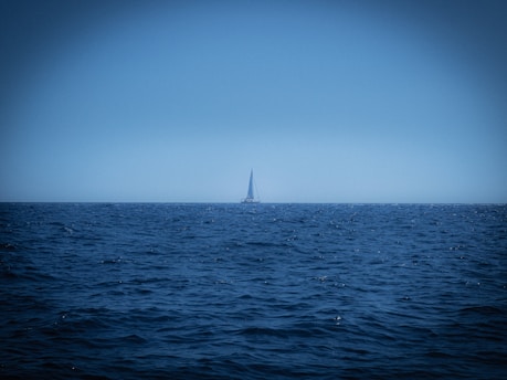 A lone sailboat cutting through deep blue ocean waves under a vast, open sky at sunset.