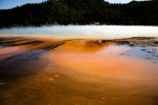 A stunning geothermal landscape with vibrant orange, yellow, and brown mineral deposits on the ground, surrounded by blue and white steam or fog. The background features a dark green forested mountain, creating a striking contrast in colors.