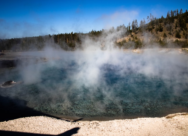 A misty geothermal pool with steam rising from the surface, surrounded by forested hills. The water appears deep blue and is bordered by a rocky shoreline. Trees in the background hint at a natural, serene setting.