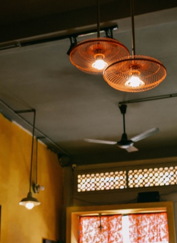 A close-up of a ceiling in a room featuring several hanging lights with intricate, circular lampshades. The ceiling also has a ceiling fan and a small section of a decorative lattice with light filtering through. The walls are painted a warm shade.