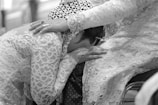 A person adorned in elaborate lace attire rests their head on the lap of another, who gently places a hand on their back. The scene conveys intimacy and emotional support through the intricate detailing of the garments and the tender gesture.