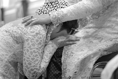 A person adorned in elaborate lace attire rests their head on the lap of another, who gently places a hand on their back. The scene conveys intimacy and emotional support through the intricate detailing of the garments and the tender gesture.