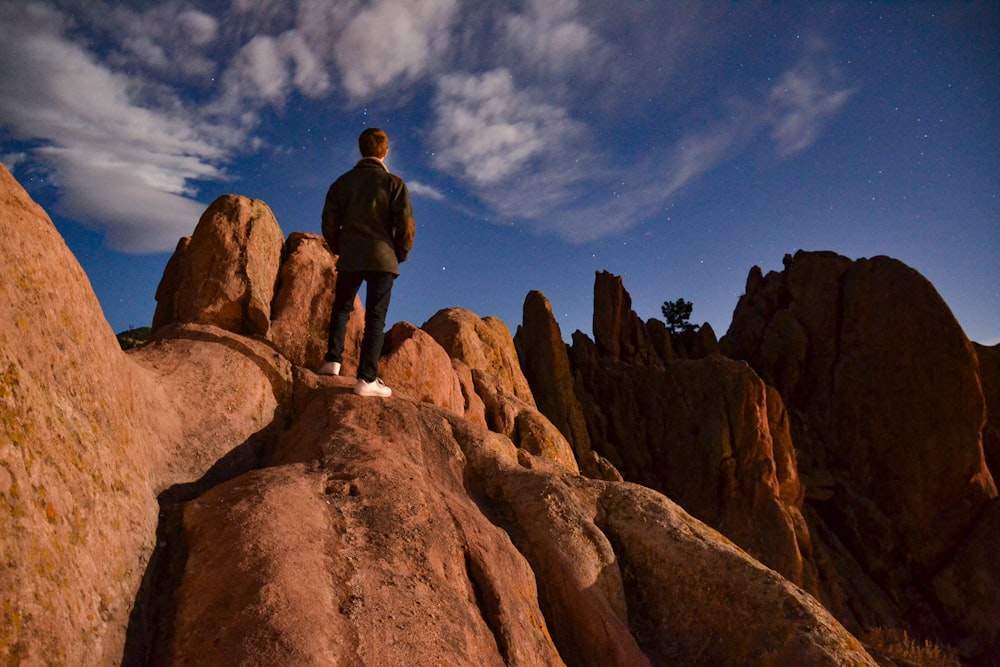 man in black jacket standing on brown rock formation under blue sky and white clouds during