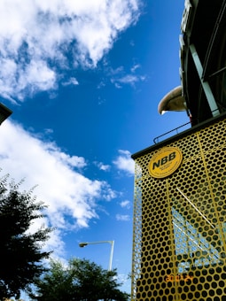 A bright blue sky with scattered white clouds stretches above a tall structure covered in a yellow perforated facade. The structure features a circular logo with the letters 'NBB' and a slogan. In the foreground, leafy trees partially obscure a streetlamp.