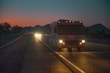 A large vehicle, possibly a recreational vehicle, travels down a dimly lit road at dusk or dawn. The sky displays hues of orange and blue, indicating sunrise or sunset. Car headlights in the distance are visible, and the silhouette of mountains can be seen in the background.