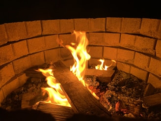 A circular stone fire pit contains burning wooden logs. Bright orange flames flicker upwards, casting a warm glow against the neatly arranged bricks that form the structure of the pit.