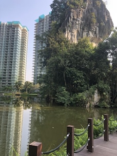 Tall residential buildings rise above a serene lake surrounded by lush greenery and trees. A rocky cliff face dominates the scene, adding a natural element to the urban setting. A wooden boardwalk with chain railing borders the lake.