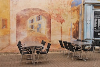 A mural covers the wall, depicting an arched passageway leading to buildings with windows. In the foreground, several chairs are arranged around two tables on a cobblestone surface. The mural incorporates warm colors and artistic details that blend with the real-life setting.