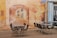 A mural covers the wall, depicting an arched passageway leading to buildings with windows. In the foreground, several chairs are arranged around two tables on a cobblestone surface. The mural incorporates warm colors and artistic details that blend with the real-life setting.
