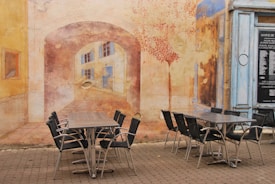 A mural covers the wall, depicting an arched passageway leading to buildings with windows. In the foreground, several chairs are arranged around two tables on a cobblestone surface. The mural incorporates warm colors and artistic details that blend with the real-life setting.