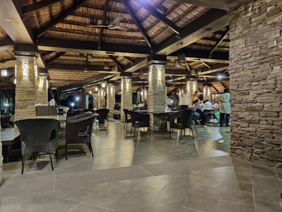 A well-lit restaurant with rustic wooden ceilings and stone pillars. There are multiple tables and chairs, some of which are empty while others are occupied by people dining. The ambiance appears cozy and inviting, with warm lighting fixtures hanging from the ceiling.